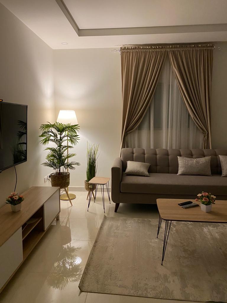A modern living area is presented with a cozy sofa and a stylish coffee table. A flat-screen TV is mounted on the wall, complemented by soft lighting from a nearby floor lamp. Decorative plants and small accent tables add a touch of warmth to the space.