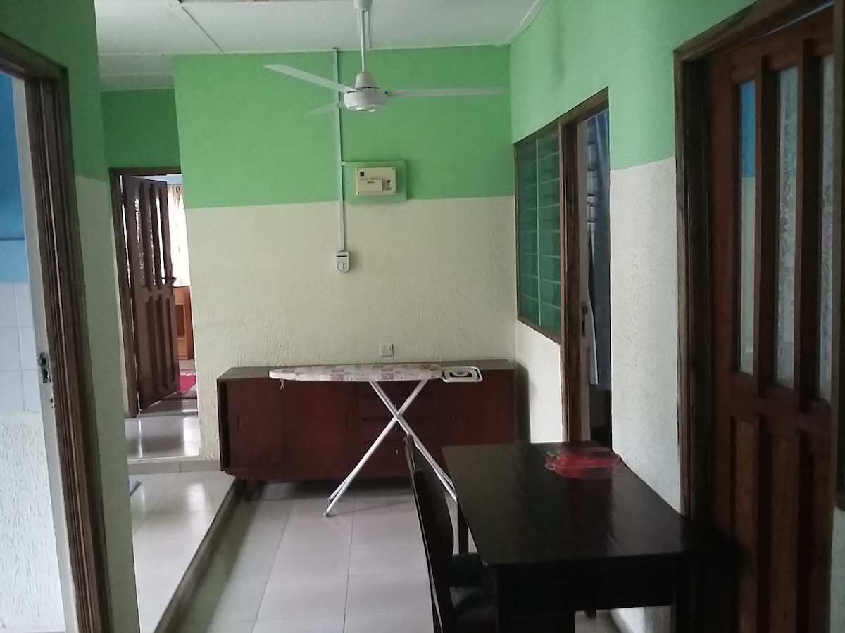 A hallway is shown, featuring green and white painted walls. A wooden table with a red plate is positioned beside a wardrobe. An iron board is set up against the wall. Ceiling fans are visible, contributing to the airy environment.