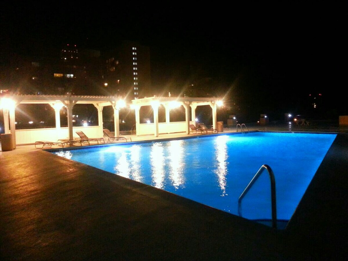 The pool area is illuminated by soft lighting, creating a serene atmosphere. The spacious pool features a striking blue color reflection, surrounded by lounge chairs. A covered area adjacent to the pool offers shade and seating, highlighting the inviting outdoor space.