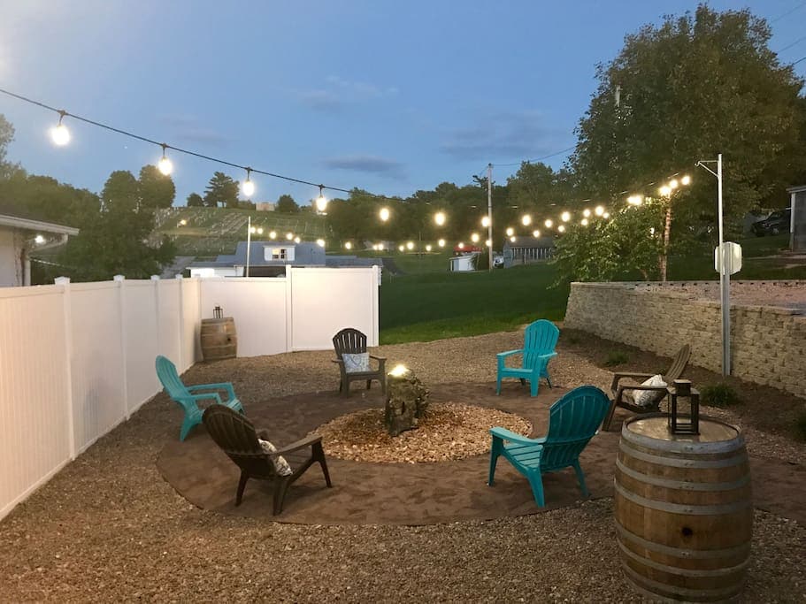 An outdoor seating area is surrounded by a low fence, featuring a circular arrangement of chairs and a fire pit at the center. Softly illuminated by string lights overhead, the space offers a gravel base with wooden barrels placed alongside.