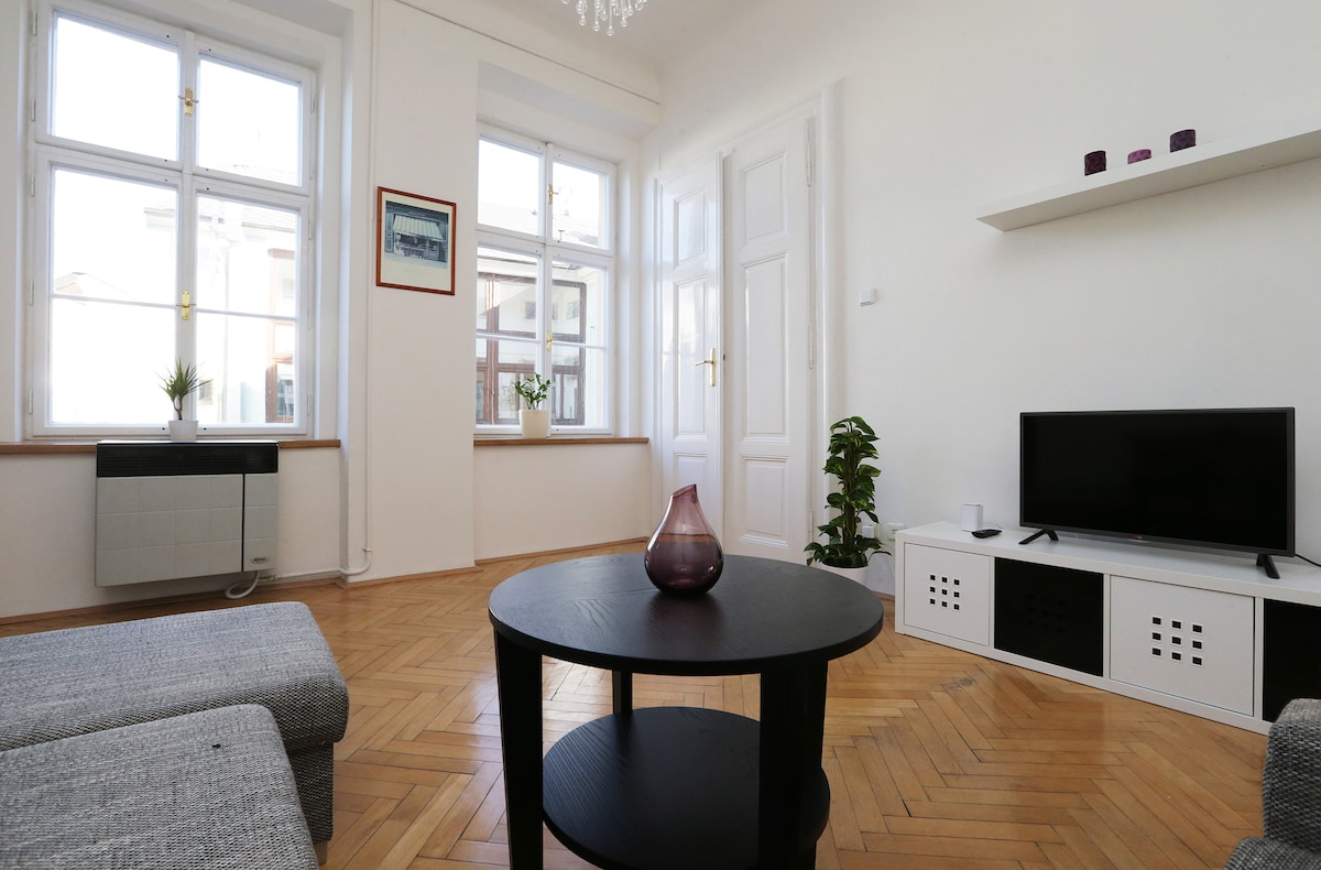 A bright living area features large windows allowing natural light to fill the space. A round black coffee table sits in the center, accompanied by a gray sofa. A wall-mounted television and decorative items are positioned on a low shelf, enhancing the room's simplicity.