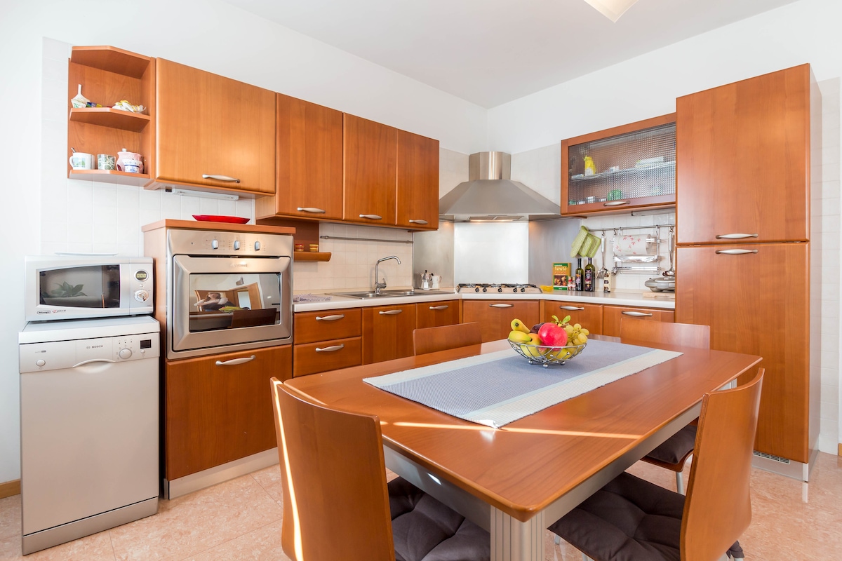 The kitchen and dining area features wooden cabinetry with modern appliances, including a gas stove, oven, microwave, and dishwasher. A dining table is set with a bowl of fresh fruit, while natural light illuminates the space through the window.