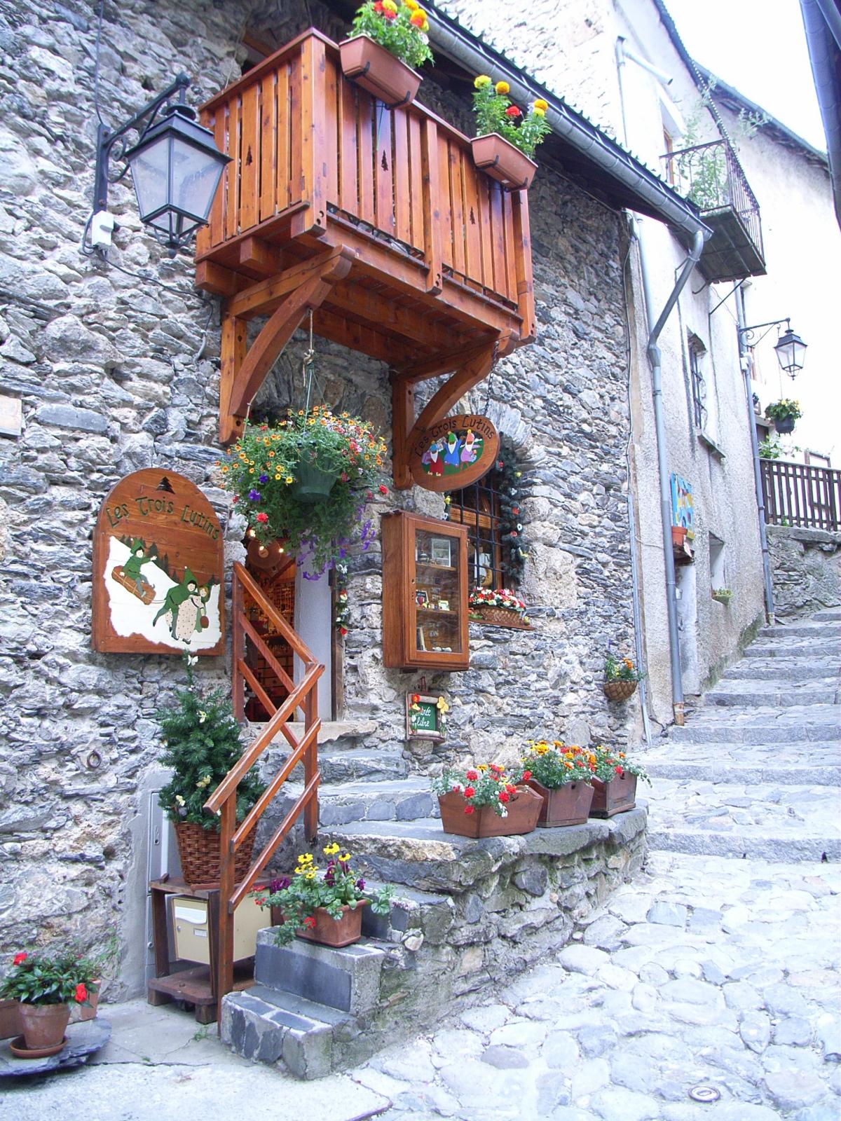 A charming entrance features a wooden balcony adorned with potted flowers, complemented by hanging planters. The rustic stone wall and cobblestone pathway lead to a welcoming shopfront, framed by colorful floral arrangements at the doorstep.