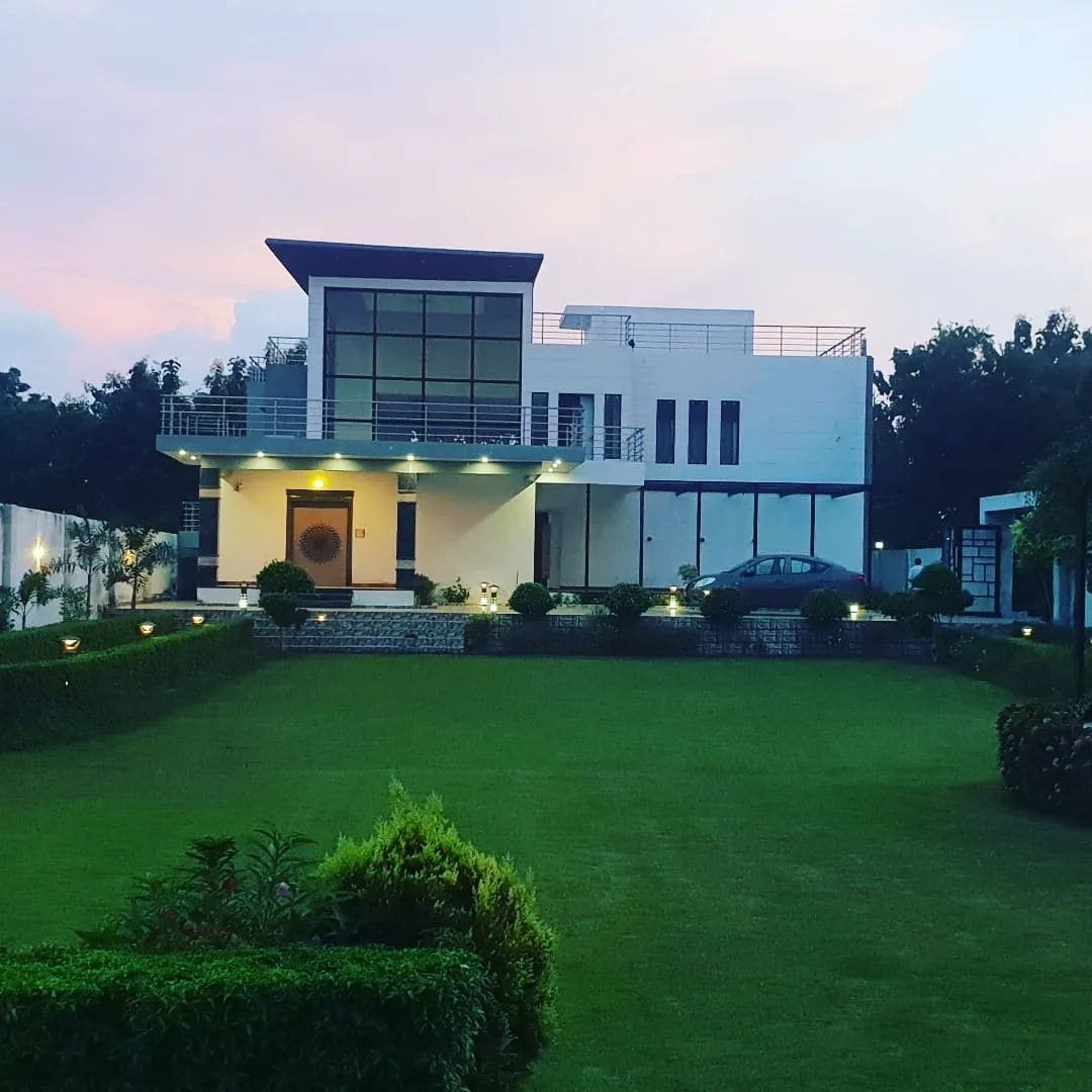 A modern villa is set against a vibrant sunset sky, showcasing large glass windows and a spacious balcony. The well-maintained lawn is surrounded by neat hedges and is illuminated by soft landscape lighting, creating a serene environment.