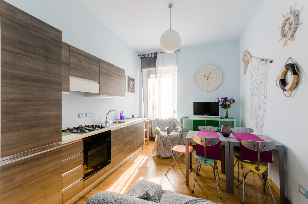 A modern kitchen area is contrasted with vibrant furniture, including a colorful dining table and chairs. Natural light fills the space through a window, and a soft seating arrangement invites relaxation. Decorative elements include a wall clock and a small television.