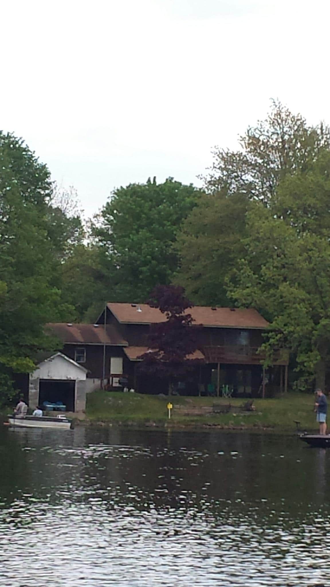 A serene lakeside view features a two-story house surrounded by lush greenery. A small boathouse stands to the side, with two individuals engaged in activities on the water. The landscape is enveloped in shades of green, reflecting the tranquility of the setting.