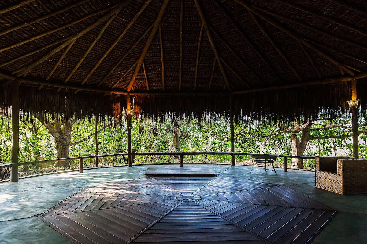 A spacious open pavilion features a wooden floor with circular designs, illuminated by soft lighting. Natural materials used in the thatched roof create a harmonious connection to the surrounding trees, enhancing the serene environment ideal for relaxation and yoga.