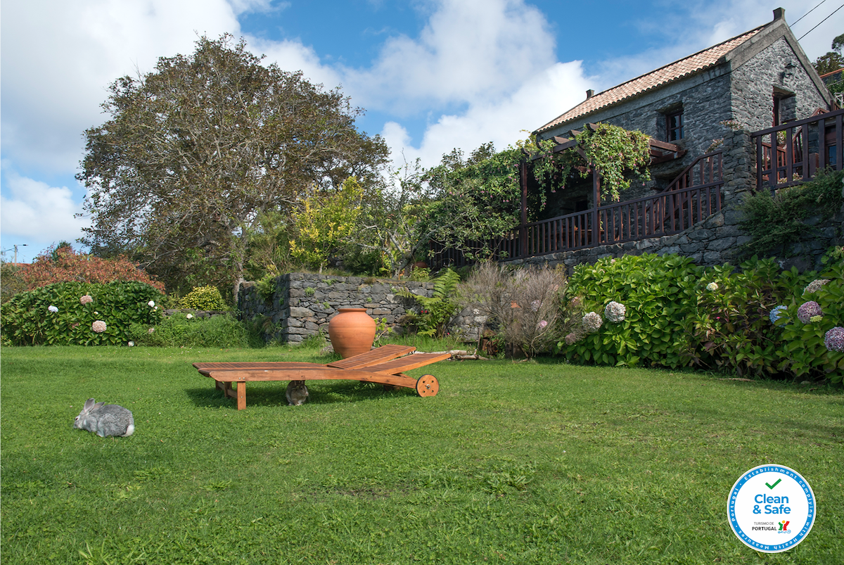 A lush green garden surrounds a stone building, showcasing a wooden seating area with an earthen pot. Two rabbits are seen on the grass, and colorful flowers frame the space, contributing to a natural and tranquil atmosphere.