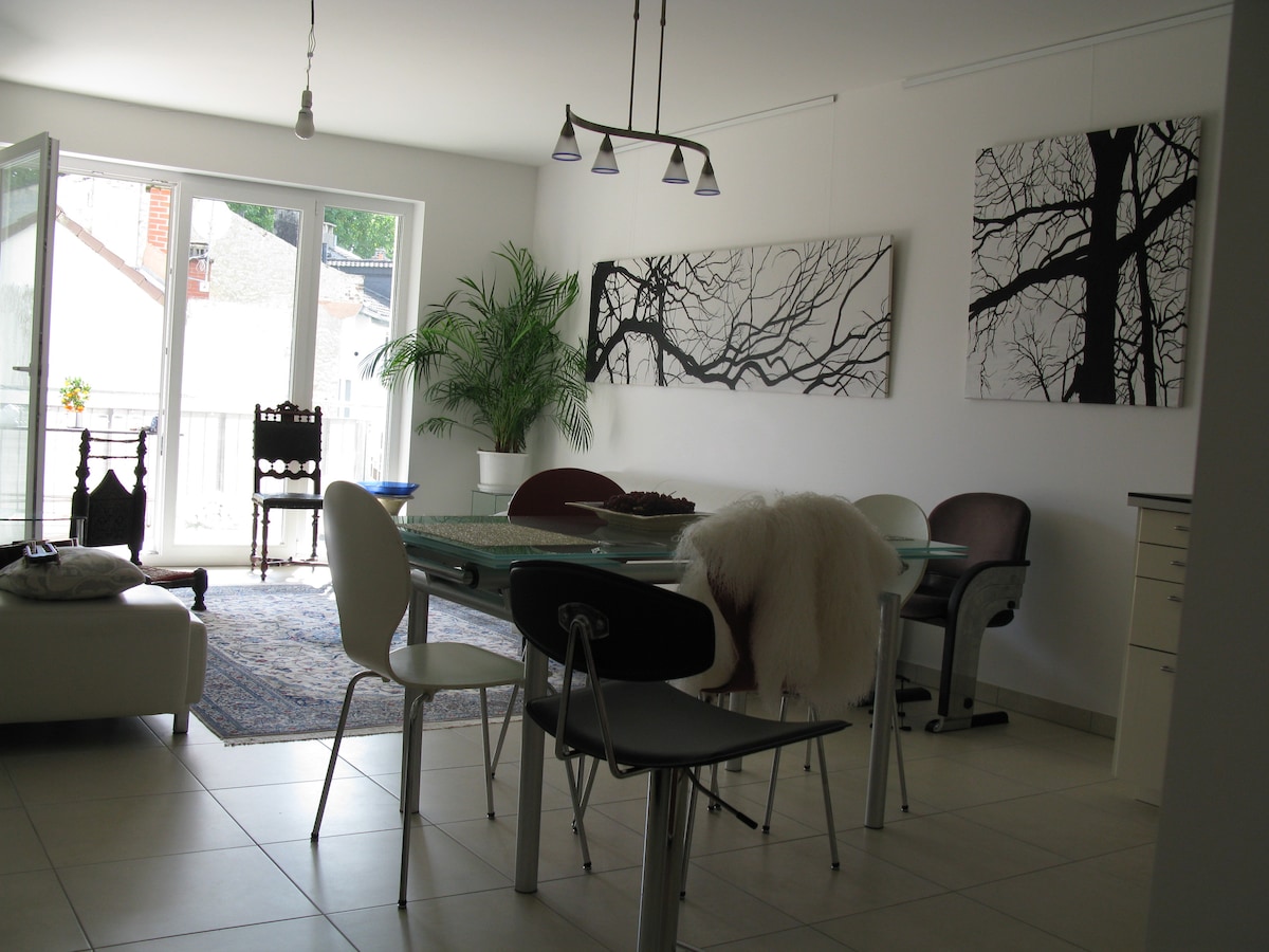 A spacious living area features a large dining table surrounded by various chairs, including a white sofa. Large windows allow natural light to fill the room, complemented by modern artwork depicting tree branches. A rug adds texture, and a potted plant enhances the atmosphere.