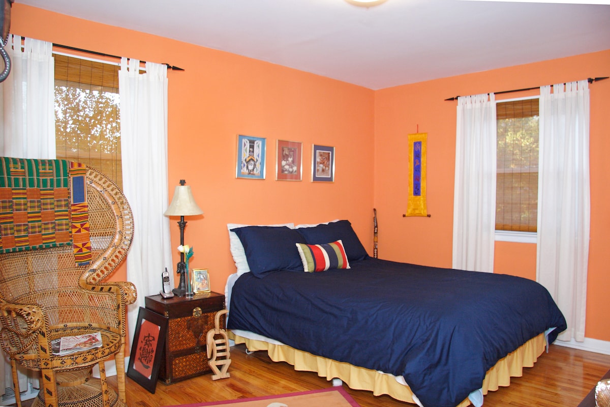 A spacious bedroom features an orange accent wall complemented by warm wooden flooring. A comfortable bed, adorned with dark bedding and decorative pillows, is flanked by two windows with bamboo shades. A cozy chair and nightstand provide additional furnishings, contributing to a welcoming atmosphere.