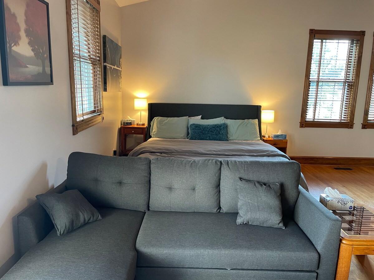 A spacious room features a comfortable king-size bed with two bedside lamps providing soft light. In the foreground, a gray sectional sofa offers seating, complementing the room's inviting layout. Natural light streams through the blinds of two windows, enhancing the serene atmosphere.
