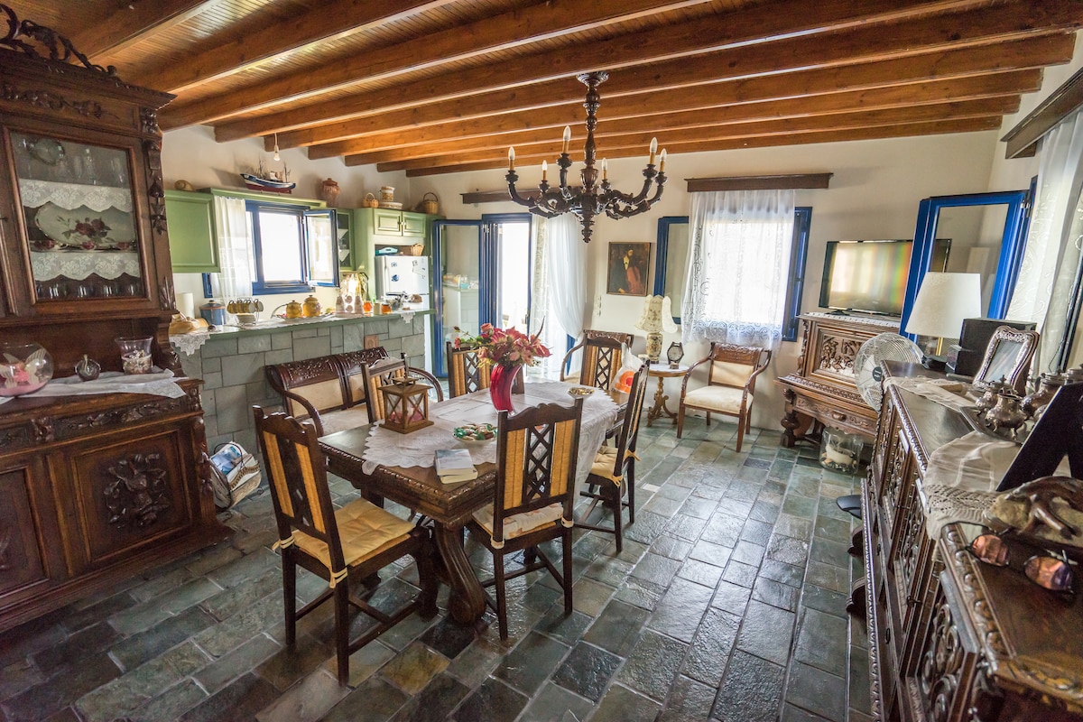 A spacious dining area is presented with a large wooden table surrounded by six chairs. A classic chandelier hangs overhead, while a wall cabinet displays decorative items. Light streams in through two windowed doors, illuminating the natural stone floor.
