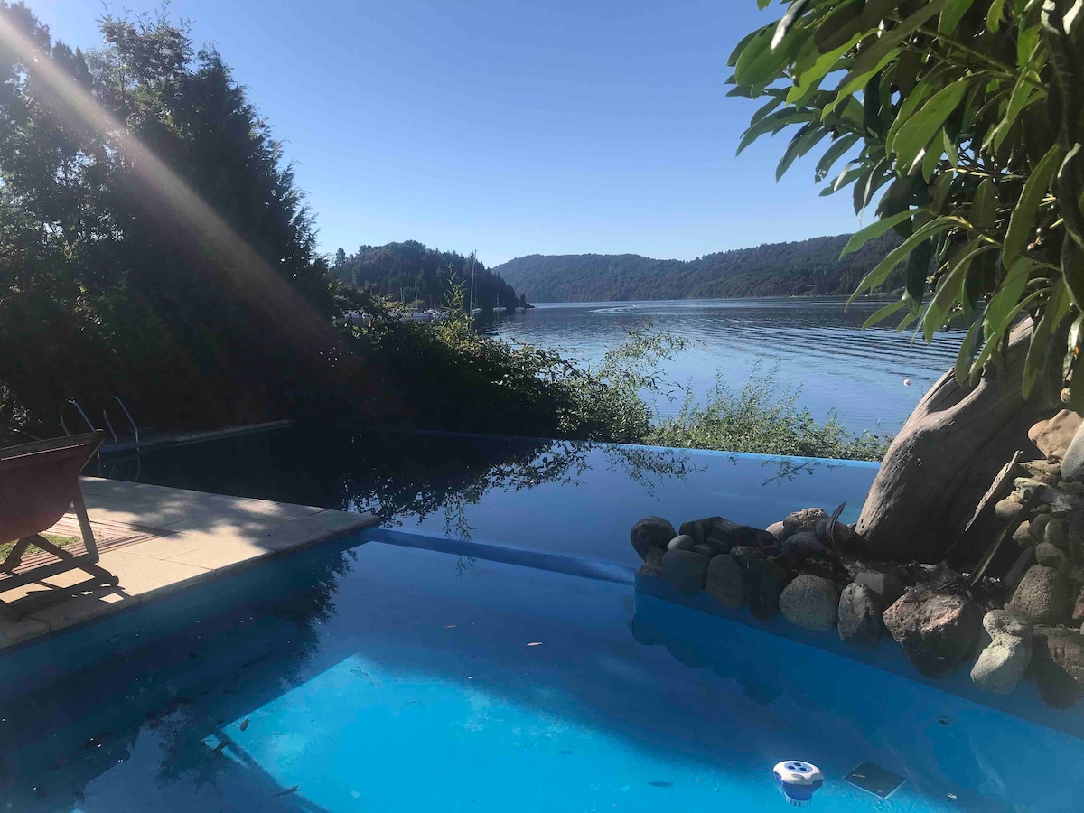 An outdoor swimming pool extends towards a serene lake, surrounded by lush greenery. Natural rock features enhance the pool area, with a sunbed positioned nearby. The calm water reflects the clear blue sky and distant hills, creating a tranquil view for guests.
