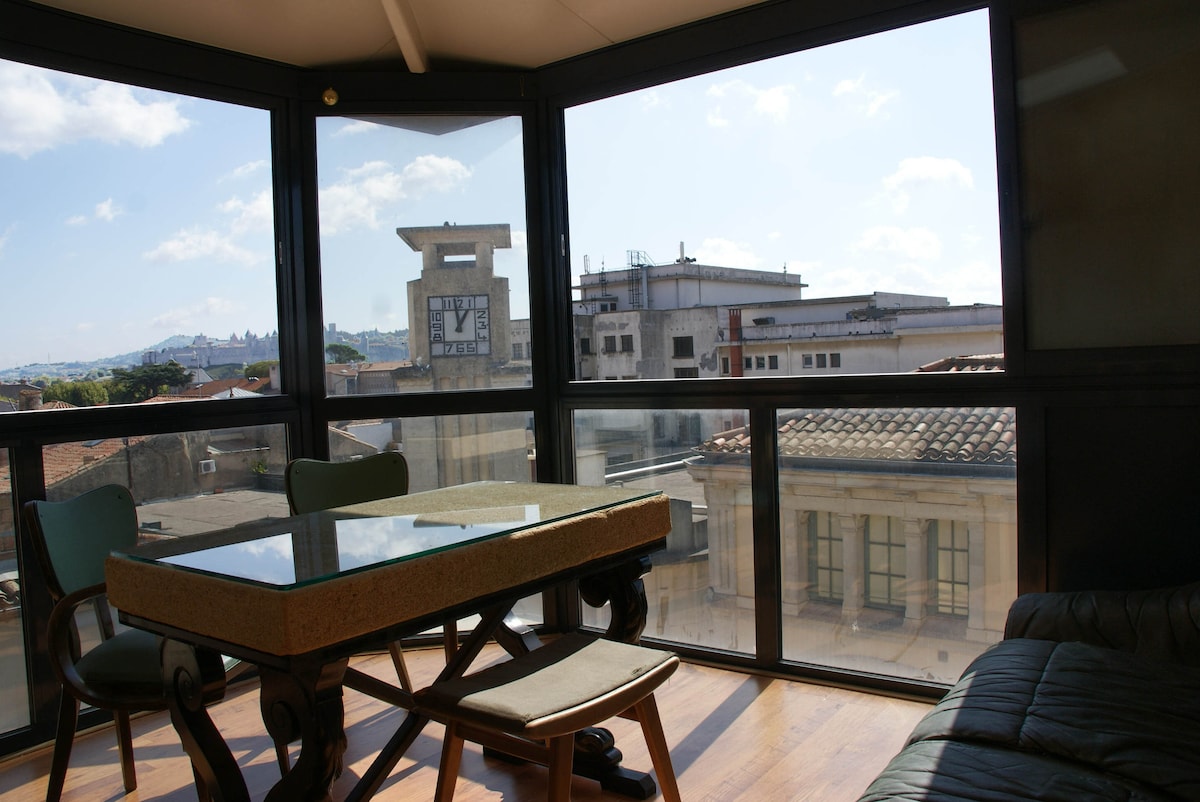 A light-filled veranda features floor-to-ceiling glass walls, showcasing views of the surrounding rooftops and the iconic medieval city in the distance. A sleek table is positioned at the center, accompanied by two chairs, offering a perfect setting for meals or relaxation.