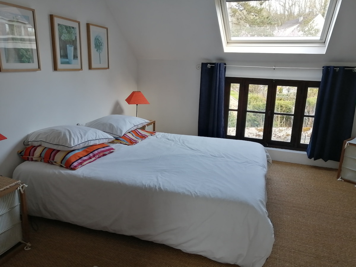 A bright bedroom features a spacious double bed with white linens and colorful pillows. Two nightstands with lamps are positioned beside the bed. A large window brings in natural light, overlooking a garden, and framed artwork adorns the walls.