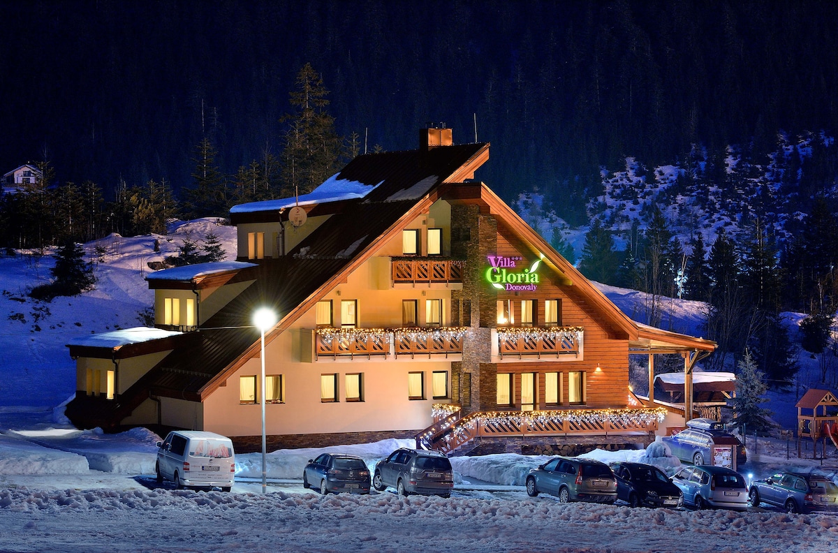 The lodge is illuminated at night, showcasing its wooden architecture and expansive balconies. Snow covers the ground, while several vehicles are parked in front. Tall trees surround the property, contributing to a serene winter atmosphere.