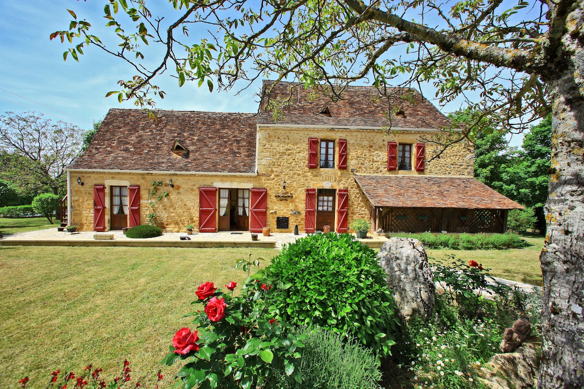 A charming 18th-century stone house is framed by vibrant greenery and colorful flowerbeds. Red shutters accent the exterior, and a spacious lawn extends around the property. The surrounding trees provide a serene atmosphere, reflecting the home's inviting and tranquil setting.