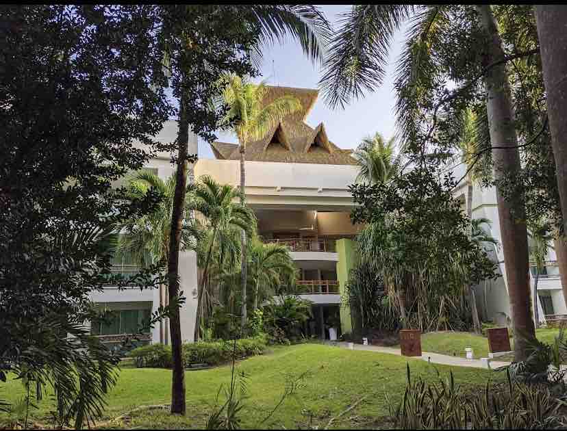 The exterior of an elegant building is framed by lush greenery and palm trees, creating a serene atmosphere. Architectural details, including a thatched roof, add character. The well-maintained lawn complements the tropical setting, enhancing the overall appeal of the complex.