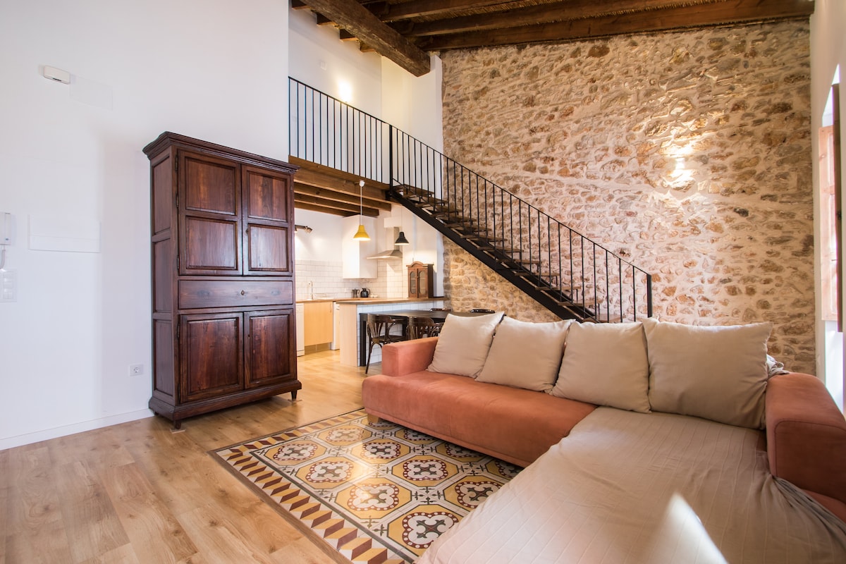 An open and bright living space features a large, comfortable sofa set against a textured stone wall. A wooden cabinet adds richness to the room. A staircase leads to an upper area, with natural light enhancing the inviting ambiance.