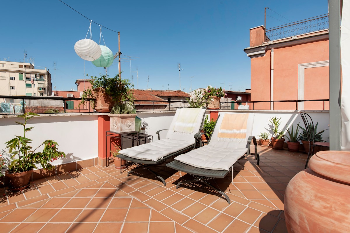 A shared rooftop terrace is depicted, featuring two lounge chairs positioned on a tiled surface. Potted plants are arranged along the perimeter, while colorful lanterns hang above. A clear blue sky creates a bright backdrop, enhancing the inviting outdoor space.