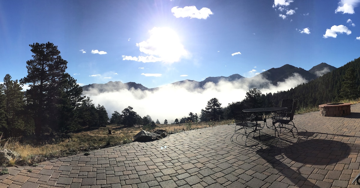 A stone-paved outdoor area is illustrated, with a metal table and chairs positioned to face views of distant mountains and a cloud-filled valley below. The scene is enhanced by bright sunlight streaming from a clear blue sky, with a few scattered clouds above.
