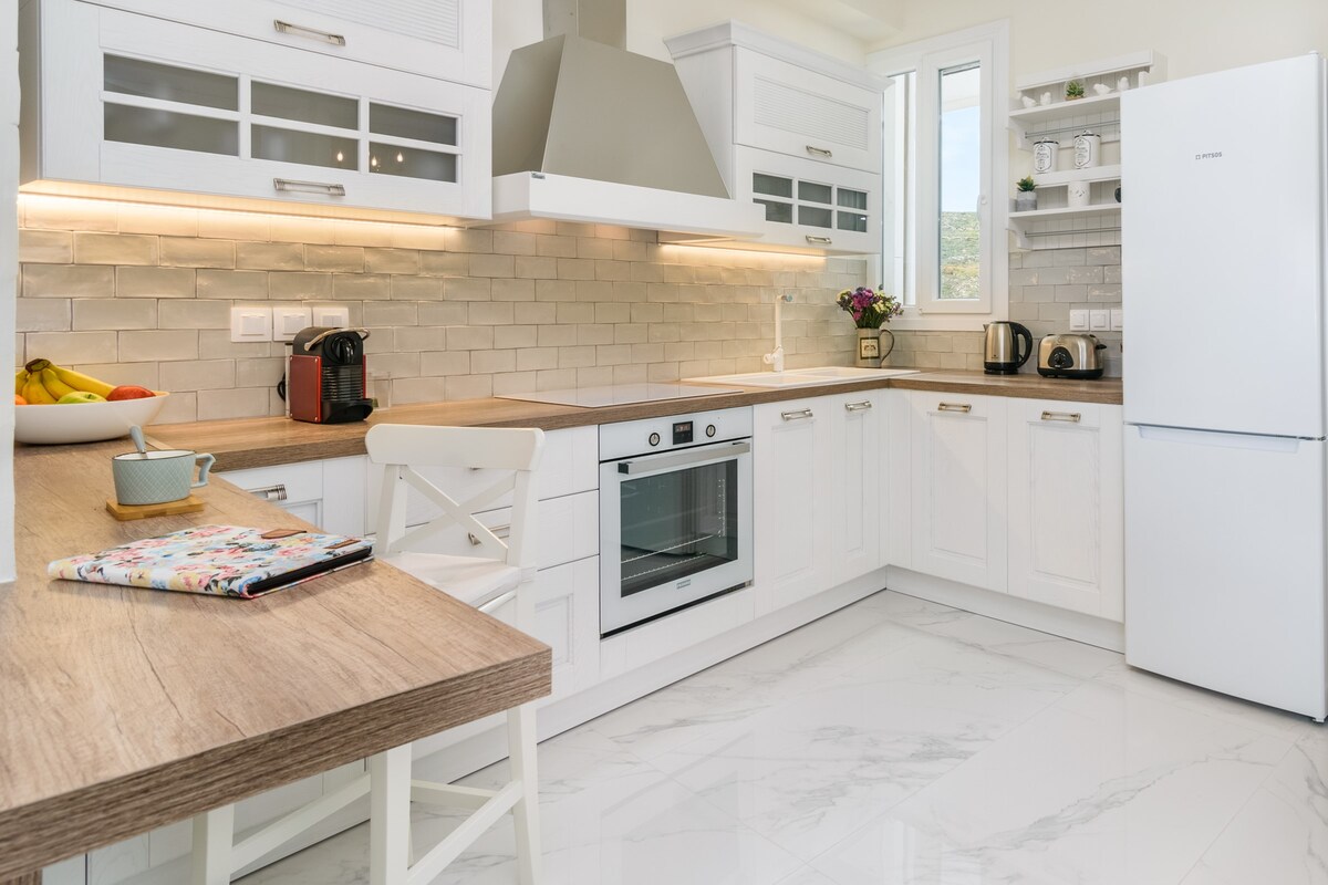 A modern kitchen features white cabinetry with glass accents and a light-colored countertop. Essential appliances, including an oven and refrigerator, are visible. A small table holds a colorful notepad and a mug, while fresh fruit is displayed. Natural light enhances the inviting space.