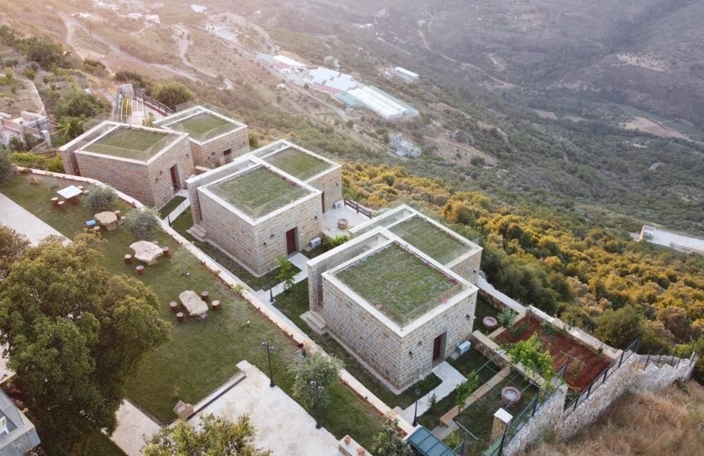 Aerial view of eco-friendly stone suites with green roofs surrounded by lush gardens. A serene landscape is visible in the background, featuring rolling hills and farmland. Outdoor seating areas with picnic tables are included, emphasizing the connection with nature.