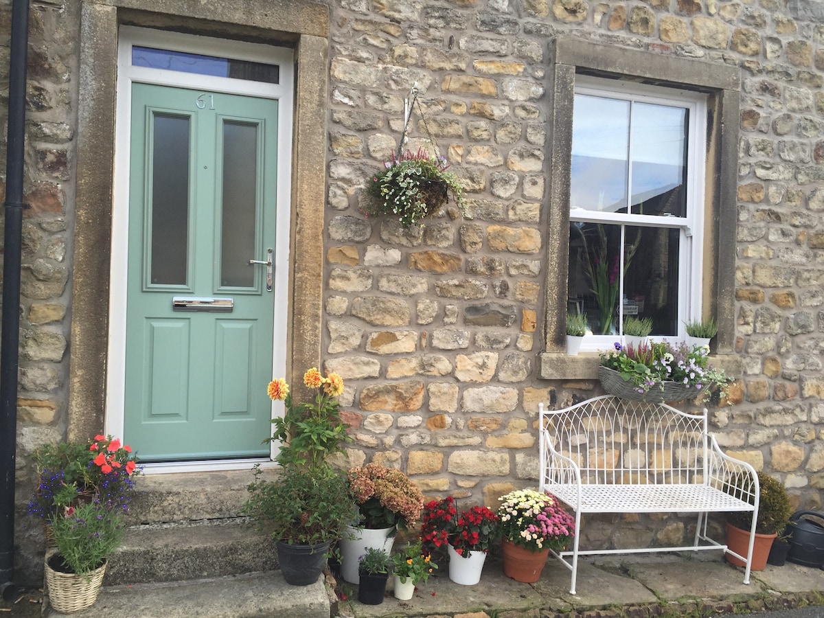 A charming entrance is presented, featuring a light green door set within a stone facade. The door is flanked by a window adorned with potted flowers. A white bench sits in front, surrounded by vibrant floral arrangements in pots, enhancing the welcoming appearance.
