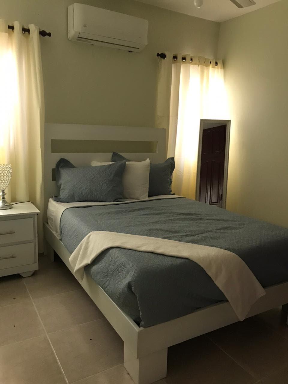 A cozy sleeping area features a spacious bed with soft blue and white bedding. Adjacent to the bed, a nightstand holds a decorative lamp. Natural light filters through light-colored curtains, and an air conditioning unit is mounted above the bed.