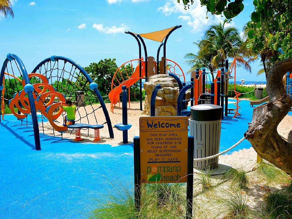 A vibrant playground is showcased, featuring unique climbing structures and slides designed for children. Brightly colored equipment in orange and blue contrasts with the soft rubberized floor. Lush green palm trees and a clear blue sky add to the cheerful atmosphere, ensuring a welcoming space for family fun.