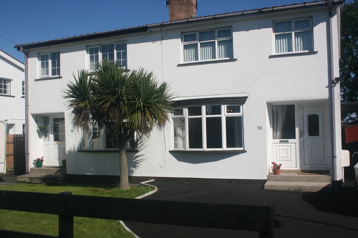 A semi-detached house is displayed, featuring a white exterior with large windows that allow ample natural light. The front yard is adorned with palm trees, adding a touch of greenery to the landscape. A black paved path leads to the entrance doors.