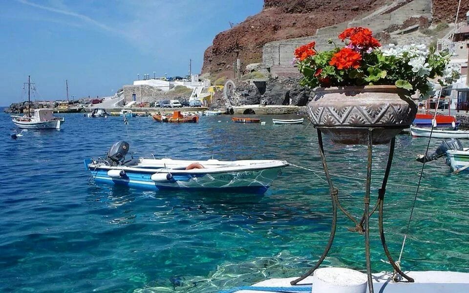 A small fishing boat is anchored in clear blue waters, complemented by vibrant flowers in a decorative pot positioned on the shore. In the background, a traditional harbor is visible, along with a range of boats gently bobbing on the waves.
