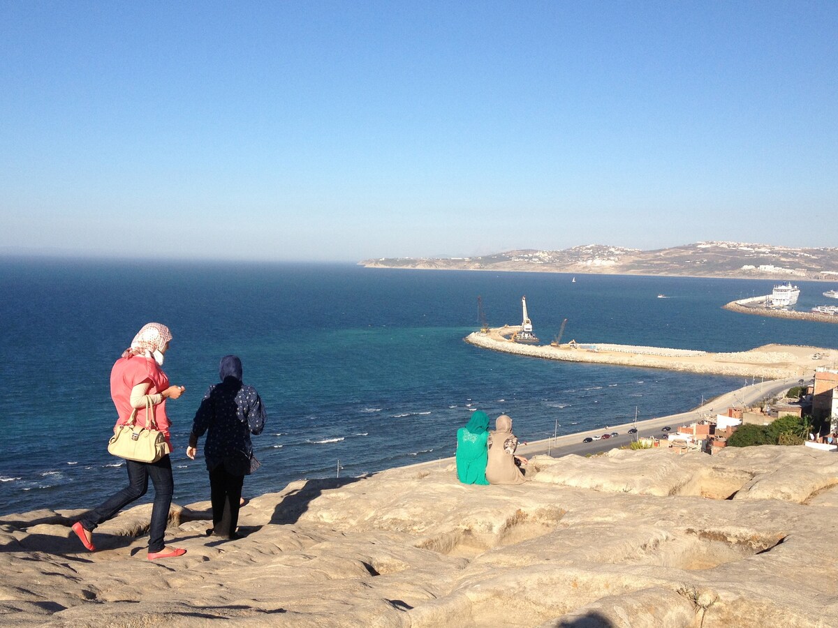 Two figures are seen walking along a rocky view point overlooking the ocean. In the distance, a harbor is visible, with boats and a lighthouse along the coastline. The clear blue sky compliments the vibrant water below.