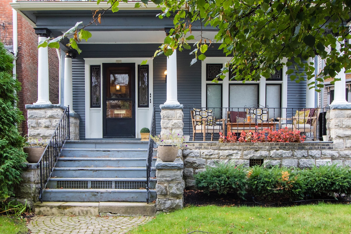 The entrance features a series of stone steps leading to a welcoming front door framed by elegant columns. A porch area with seating options is visible, surrounded by planters and vibrant greenery, creating a cozy yet inviting outdoor space.