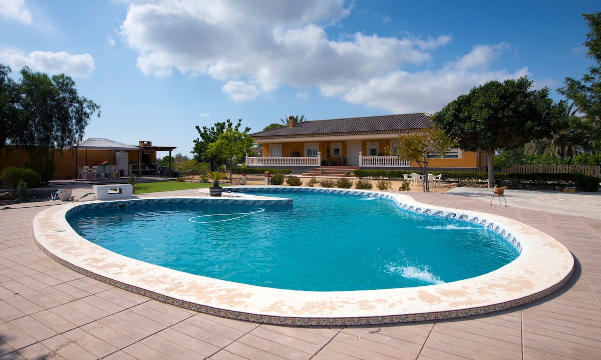 A large, inviting swimming pool is surrounded by a spacious patio. The pool features a unique shape and steps leading into the water. Lush greenery and a yellow-walled house are visible in the background, complemented by a shaded area for relaxation.