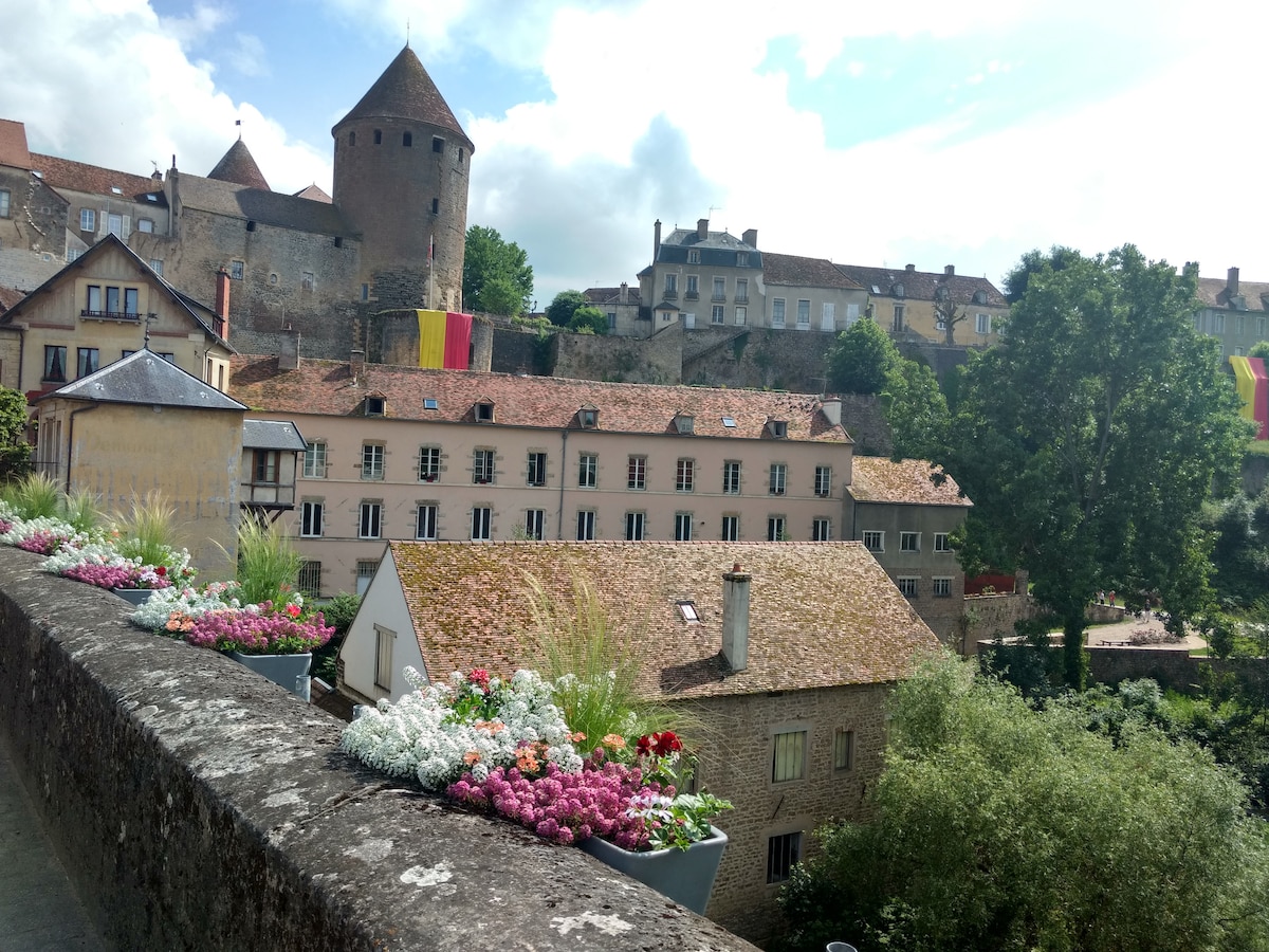 A scenic view captures traditional buildings adorned with colorful flower boxes, reflecting a charming historic atmosphere. The rooftops feature rustic tiles, with a backdrop of lush greenery and a hilltop castle, while vibrant banners add a touch of local character to the village.