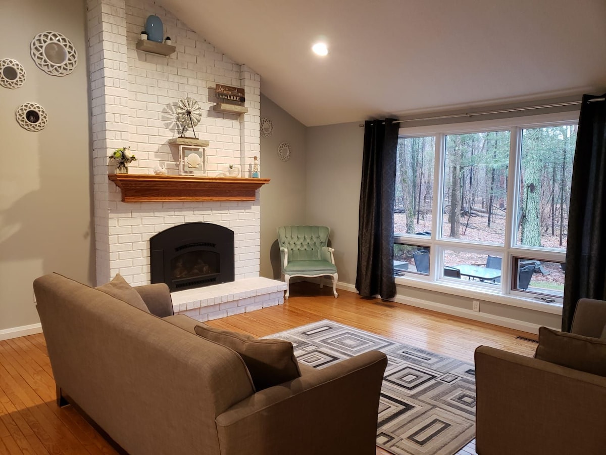 A cozy living area is highlighted by a brick fireplace with decorative accents. Large windows offer views of the surrounding forest, while comfortable seating arrangements include a light green chair and a gray sofa. A patterned area rug enhances the warmth of the space.