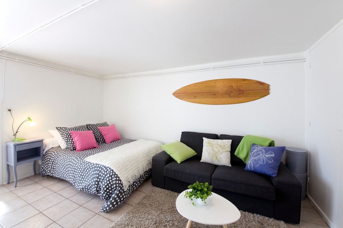 A cozy room features a spacious double bed with a textured white blanket, complemented by decorative pillows in pink and green. A dark couch with matching cushions provides an additional seating area. A wooden surfboard is mounted on the wall above, adding a coastal touch.