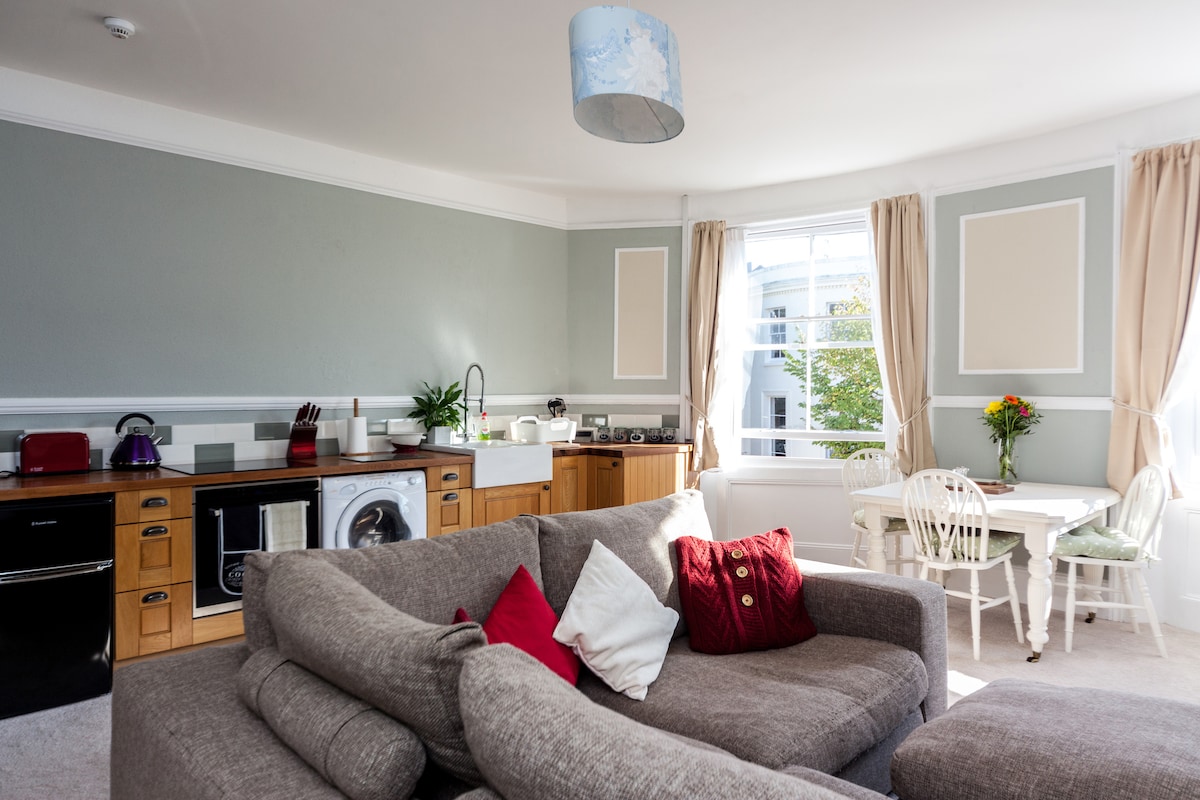 A spacious living area features a comfortable corner sofa adorned with cushions in varied colors. An adjoining kitchen is equipped with modern appliances, including an oven and microwave. Natural light floods in through large windows, illuminating a dining table set for four.