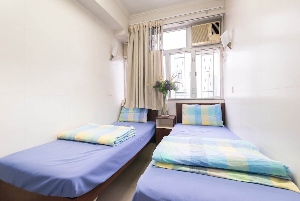 A room with two single beds is presented, featuring light blue bedding and striped throws. Natural light enters through two windows, lined with sheer curtains. A small table with a vase of flowers adds a touch of decor. An air conditioning unit is mounted on the wall.