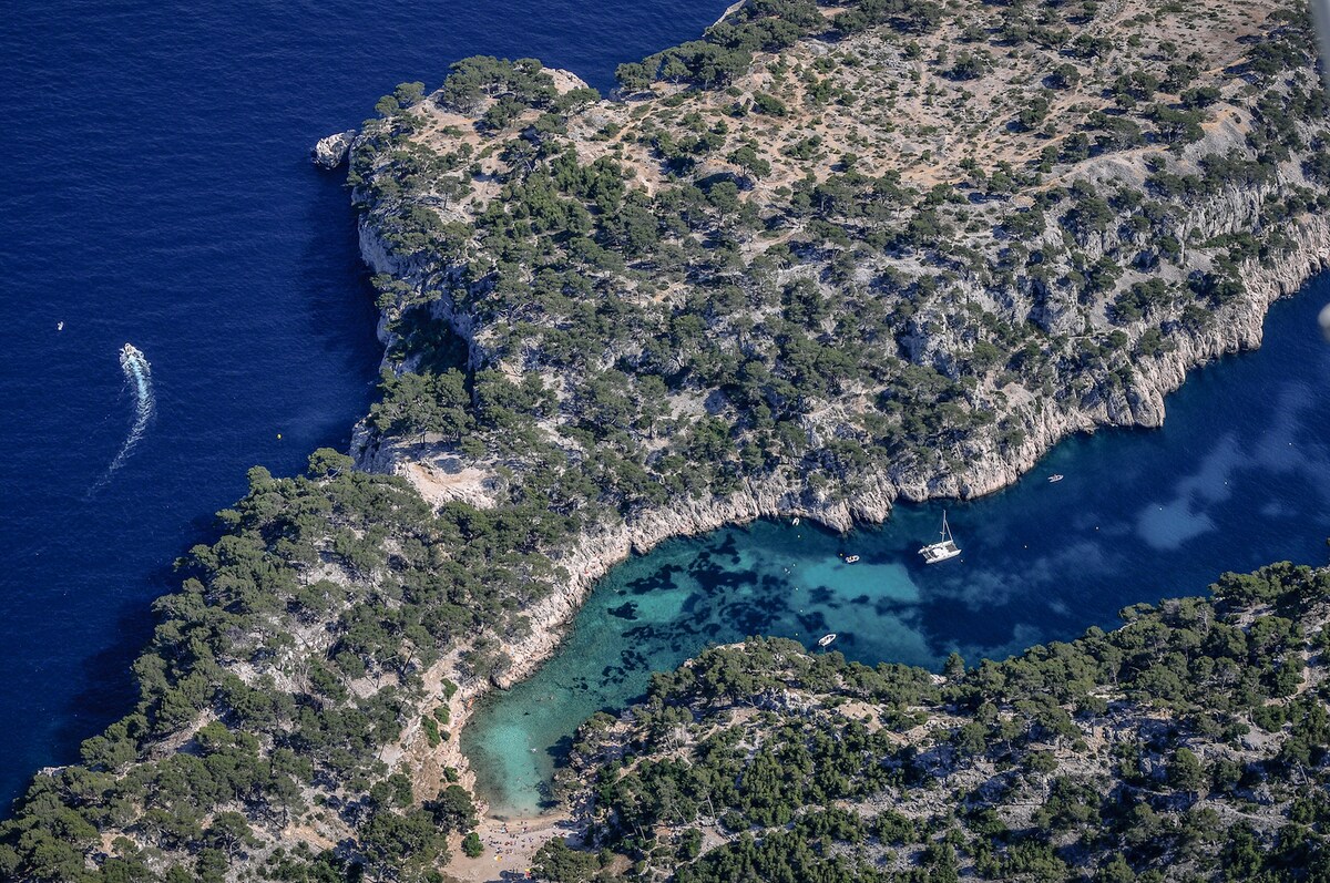 An aerial view of a secluded cove showcases clear turquoise waters surrounded by rocky terrain and greenery. Several boats are visible in the water, and the coastline features a mixture of sandy and rocky areas, inviting exploration and relaxation.