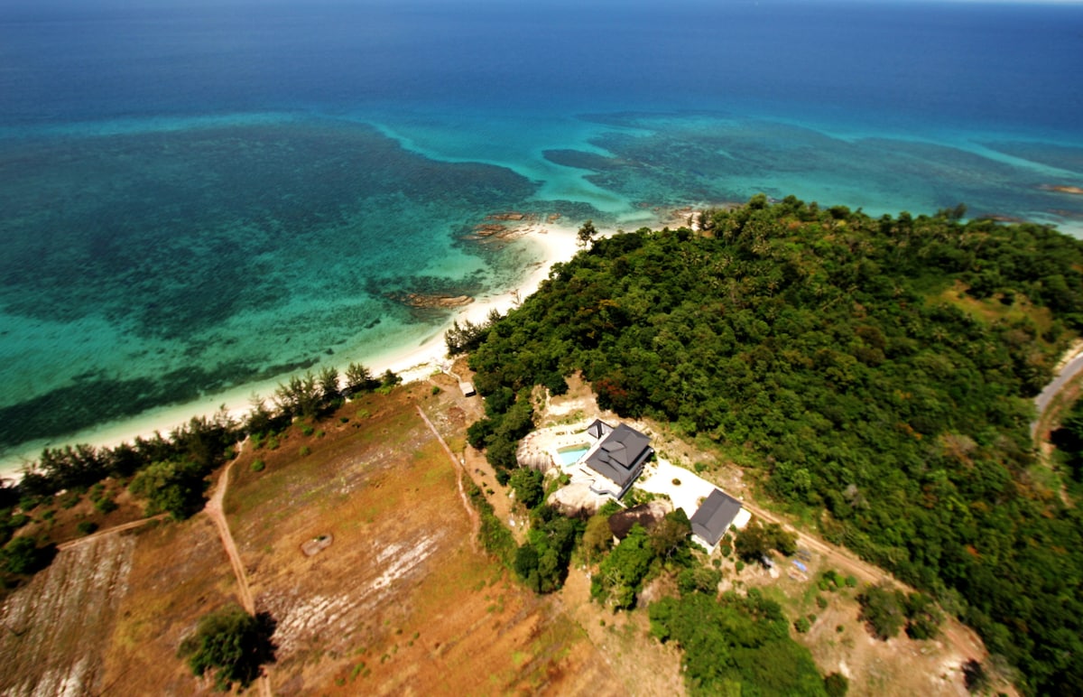 An aerial view captures the expansive coastline and lush greenery surrounding the villa. The clear turquoise waters contrast with the sandy beach, leading to vibrant coral reefs, while the villa is nestled within the tropical landscape, showcasing its private setting amidst natural beauty.