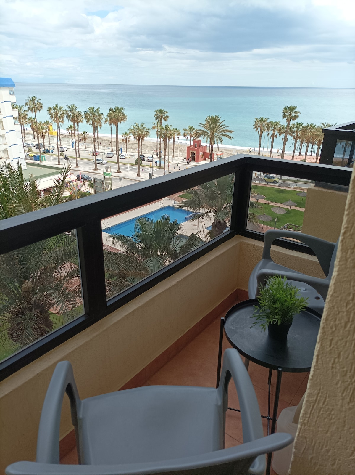A balcony offers views of the beach and the ocean, framed by palm trees. Two light-colored chairs are positioned at a small round table, which features a potted plant. The scene conveys a sense of relaxation near the water.