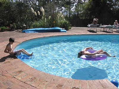 A swimming pool is surrounded by lush greenery, with the sun glistening on the water. A child sits on the edge of the pool, while another child relaxes in a floating ring. Lounge chairs are placed nearby for sunbathing.