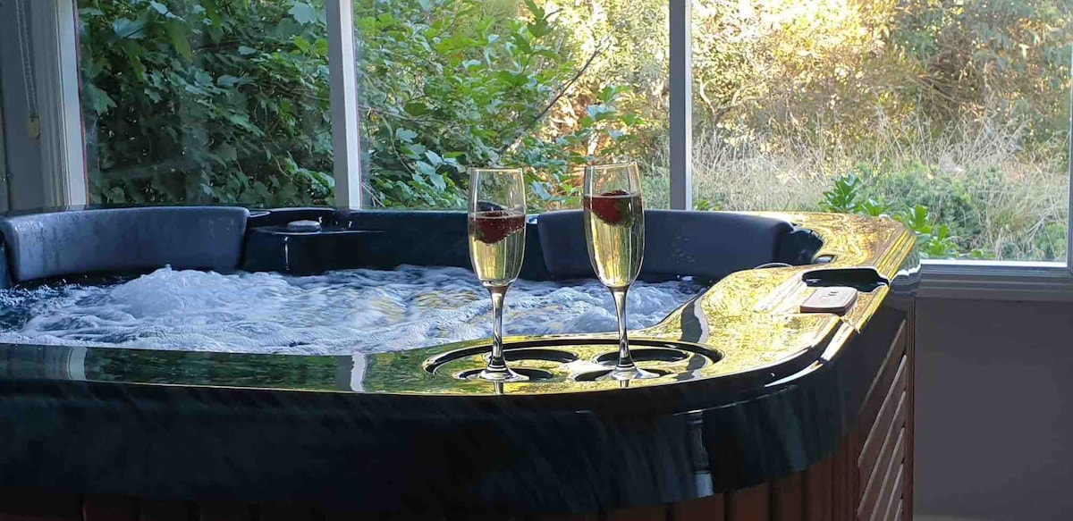 A spa featuring bubbling water is situated beside large windows, allowing natural light to flood the area. Two glasses of sparkling beverage with cherries sit on the edge, creating a serene atmosphere surrounded by lush greenery.