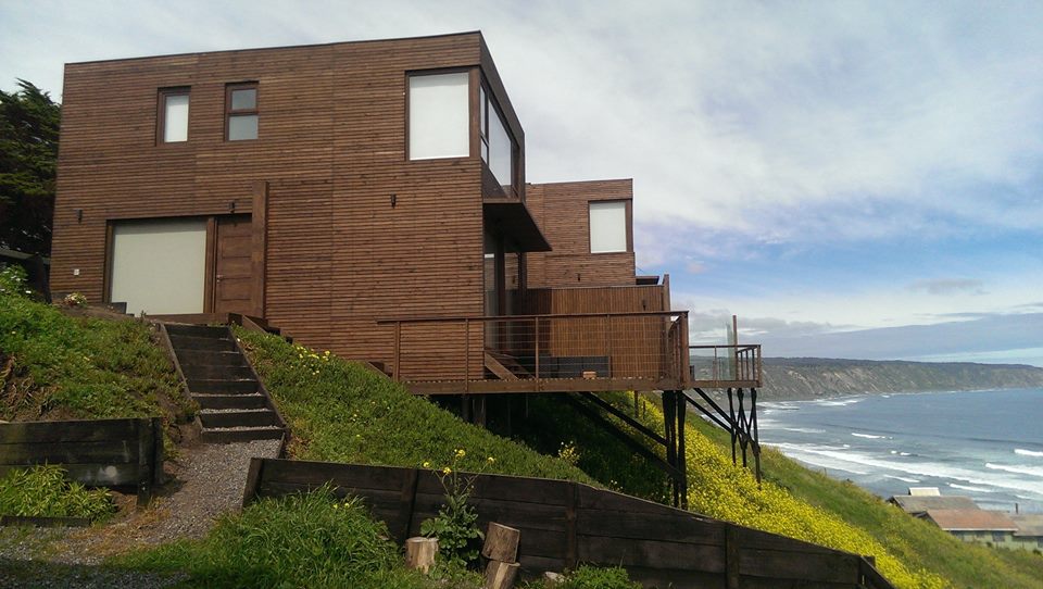A modern wooden structure is positioned on a hillside, featuring large windows that provide ocean views. The building is raised on stilts, surrounded by lush greenery and a pathway leading up to the entrance. The coastline is visible in the background, with waves gently rolling onto the shore.