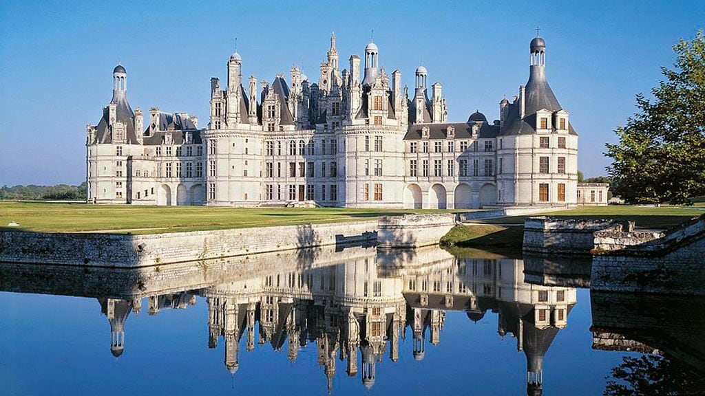A grand chateau is reflected in calm waters, showcasing its ornate architecture and multiple towers. The surrounding green landscape enhances the setting, with trees positioned near the water's edge, providing a serene view of this historic landmark.