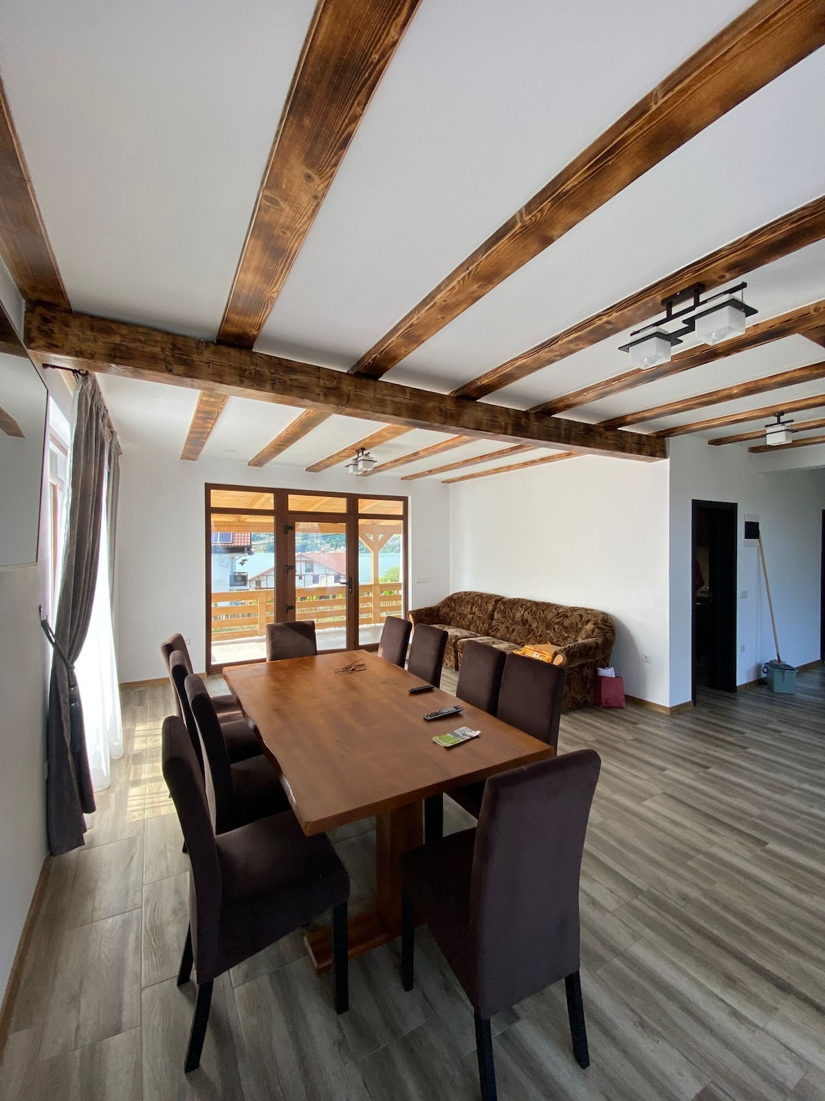 A spacious dining area is equipped with a long wooden table surrounded by eight dark chairs. Natural light enters through large windows, highlighting the beam ceiling design. A cozy sofa sits against the wall, creating an inviting space for family gatherings.