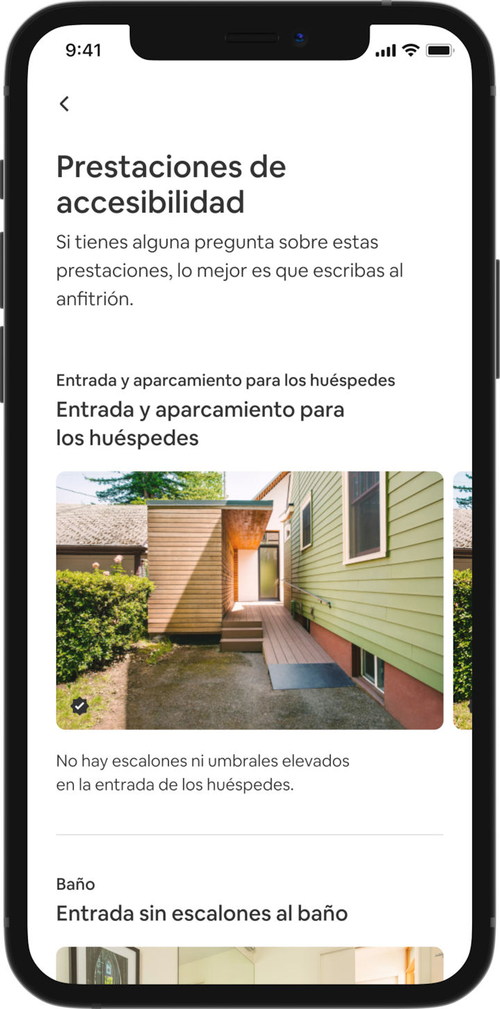 Un teléfono móvil muestra los detalles de un anuncio en Airbnb. El encabezado dice «Prestaciones de accesibilidad» y aparecen las prestaciones agrupadas por zonas, como «Entrada de los huéspedes y estacionamiento» o «Cuarto de baño». Debajo de «Entrada de los huéspedes y estacionamiento» aparece el título «Entrada sin escalones» y la descripción «No hay escalones ni umbrales elevados en la entrada de los huéspedes». Debajo aparecen varias diapositivas con fotos de las prestaciones de accesibilidad, empezando por una rampa para sillas de ruedas.