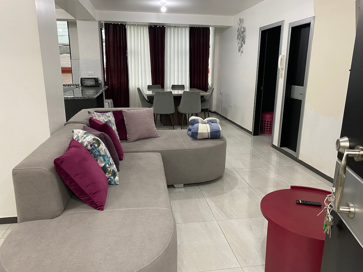 A modern living area features a grey sectional sofa with decorative pillows in purple and blue. A small round red coffee table is positioned in front, while a dining area with grey chairs is visible in the background. Large windows are dressed with dark curtains.